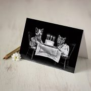 Atverama kartīte ar melnu fonu un diviem kaķīšiem pie galda