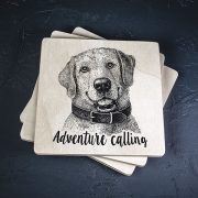Balts krūžu paliktnis ar melnu suņa zīmējumu un tekstu: "Adventure calling"