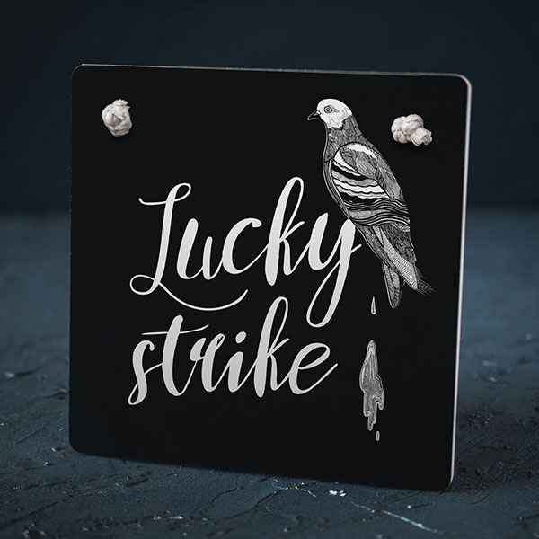 Melns dekoratīvais koka dēlītis ar baltu baloža zīmējumu un tekstu: "Lucky strike"