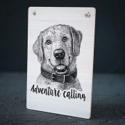 Balts dekoratīvais koka dēlītis ar melnu suņa zīmējumu un tekstu: "Adventure calling"